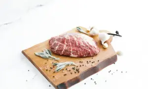 A beautiful cut of presa ibérica sits on a wooden board with sage leaves, garlic, and peppercorns, ready for a delicious meal.