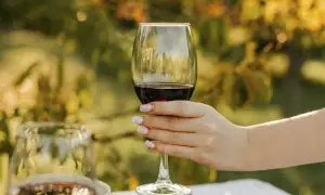A hand with decorative nails holds a glass of rich Alicante Bouschet wine, ready for a toast amidst a sun-dappled garden.