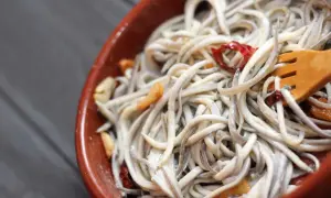 A close-up of a rustic bowl filled with a tangle of little eel, glistening with garlic and chili flakes. A wooden fork is nestled within the dish.