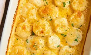 A close-up of a creamy scallop gratin in a white baking dish, topped with fresh parsley and black pepper.