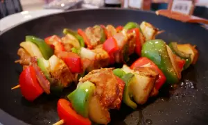Pinchos morunos sizzling in a pan, with chunks of marinated meat, red and green bell peppers, and onions on wooden skewers.