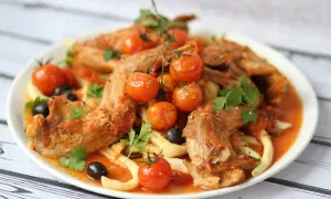A hearty rabbit stew with tender meat, cherry tomatoes, olives, and pasta, garnished with fresh parsley.