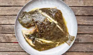 A whole grilled turbot, glistening with butter and herbs, served on a rustic plate atop a weathered wooden table.