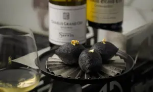A glass of chablis white wine sits beside a plate of three dark, spherical treats topped with gold leaf, with bottles of chablis grand cru in the background.