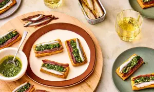 Rectangular pastries topped with red and white anchovies and green sauce. A tin of white anchovies sits nearby.