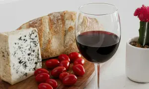 A glass of red wine, perhaps a bold trebbiano wine, sits beside a rustic loaf of bread, blue cheese, and cherry tomatoes, with a small potted cactus nearby.