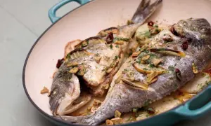 Two whole sea bream baked with lemon slices, garlic, and herbs in a teal pan, served over sliced potatoes.