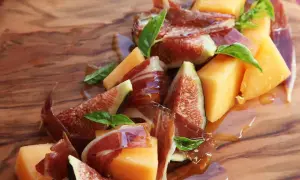 A refreshing melon con jamon appetizer with fresh figs and basil, drizzled with honey on a wooden board.