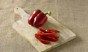 Red bell peppers, one whole and some sliced, on a wooden cutting board. Perfect for learning how to preserve peppers for later use.