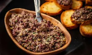 A rustic wooden bowl overflows with chunky tapenade, a savory spread of olives and herbs, ready to be enjoyed with toasted crostini.