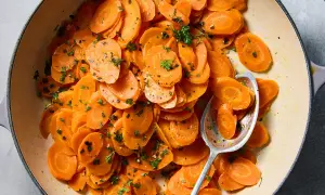 A close-up of a pot of glazed vichy carrots, glistening with butter and sprinkled with fresh parsley. A spoon rests in the pot, ready to serve.