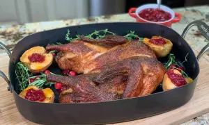 A beautifully roasted smoked duck rests in a dark roasting pan, garnished with fresh rosemary, cranberries, and baked apple halves topped with cranberry sauce.
