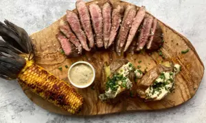 Juicy pork presa, grilled corn, and loaded baked potatoes served on a wooden board.