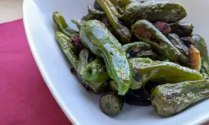 A pile of glistening, fried padron peppers in a white bowl, some with slightly charred edges, hinting at a delicious, savory snack.