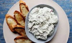 A bowl of creamy bluefish pate with herbs and red onion, served with toasted baguette slices on a light-colored plate.
