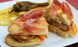 A delicious bocadillo with tender meat, cured ham, and fresh tomato, served with crispy fries and a roasted pepper.