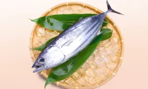 A fresh bonito, a type of tuna, rests on ice and green leaves in a woven basket.