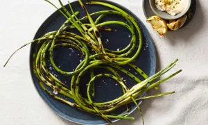 Grilled garlic scapes, seasoned with sea salt, are artfully arranged on a dark plate, with a small bowl of creamy dip and lemon wedges nearby.