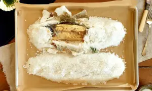 A whole salt crusted sea bass, baked to perfection, rests on a baking sheet. A lemon and herbs complete the dish.