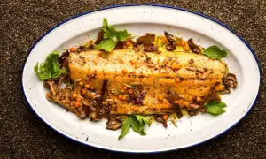 A beautifully broiled trout, glistening with olive oil and garnished with mustard seeds, sea buckthorn berries, and fresh herbs on a white enamel platter.