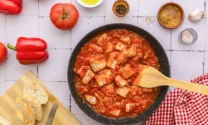 Tuna in tomato sauce simmering in a pan, with fresh tomatoes, red bell peppers, and crusty bread on the side.