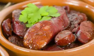 Slices of glistening chorizo al vino, simmered in a rich sauce, are served in a rustic clay bowl, garnished with fresh parsley.