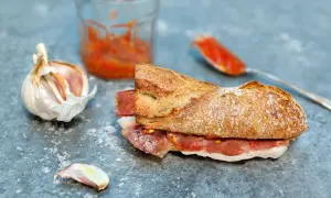 A delicious bocadillo recipe featuring a crusty baguette filled with cured ham and a vibrant tomato spread, with garlic and a jar of spread nearby.