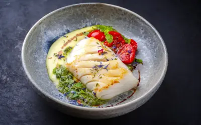 A beautifully plated poached fish recipe, featuring a white fish fillet with grilled marks, served with creamy sauce, pesto, and cherry tomatoes.