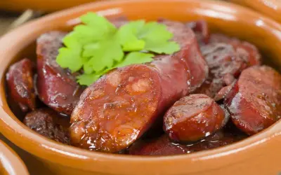 Slices of glistening chorizo al vino, simmered in a rich sauce, are served in a rustic clay bowl, garnished with fresh parsley.