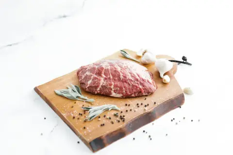 A beautiful cut of presa ibérica sits on a wooden board with sage leaves, garlic, and peppercorns, ready for a delicious meal.