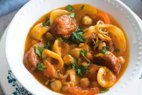 A hearty Spanish stew with tender squid rings, chickpeas, and chorizo in a rich tomato sauce, garnished with fresh parsley.
