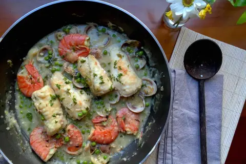 A delicious bonito fish recipe with prawns, clams, and peas in a creamy sauce, garnished with parsley.
