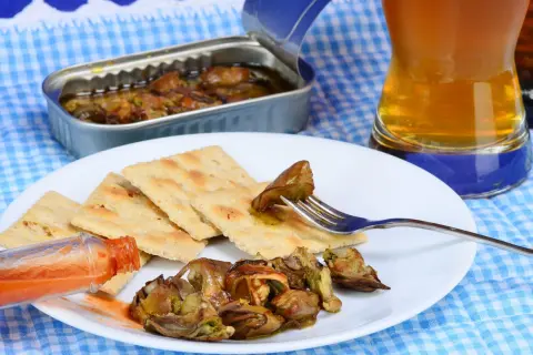 A fork lifts a smoked oyster from a plate of crackers, a bottle of hot sauce and a beer nearby. Delicious smoked oyster recipes are easy to make with these ingredients.