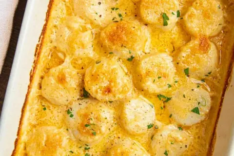 A close-up of a creamy scallop gratin in a white baking dish, topped with fresh parsley and black pepper.