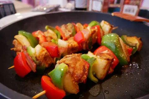 Pinchos morunos sizzling in a pan, with chunks of marinated meat, red and green bell peppers, and onions on wooden skewers.