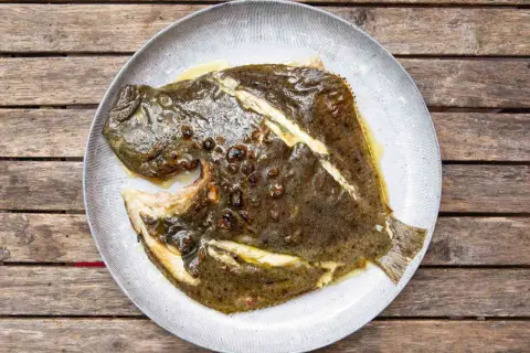 A whole grilled turbot, glistening with butter and herbs, served on a rustic plate atop a weathered wooden table.