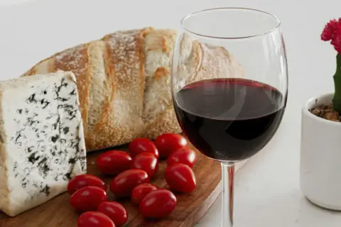 A glass of red wine, perhaps a bold trebbiano wine, sits beside a rustic loaf of bread, blue cheese, and cherry tomatoes, with a small potted cactus nearby.