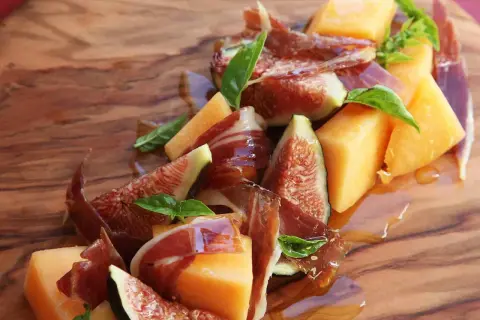 A refreshing melon con jamon appetizer with fresh figs and basil, drizzled with honey on a wooden board.