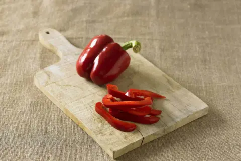 Red bell peppers, one whole and some sliced, on a wooden cutting board. Perfect for learning how to preserve peppers for later use.