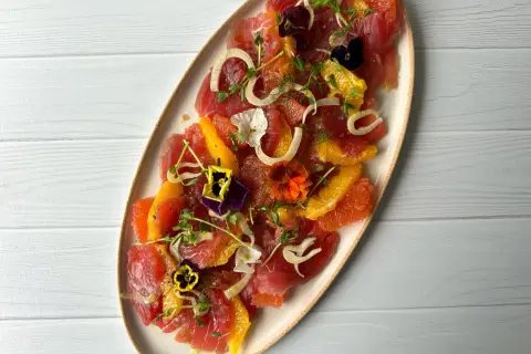 A vibrant tuna crudo dish featuring thinly sliced raw tuna, citrus segments, fennel, microgreens, and edible flowers.