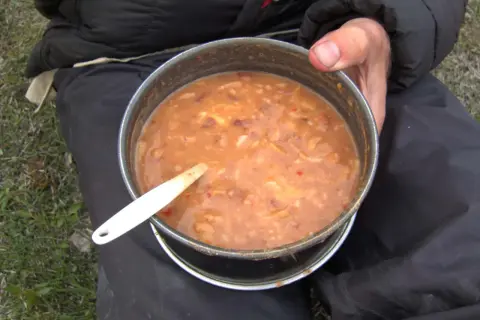 A hearty bowl of gulas fish stew, served in a metal pot with a white spoon, ready for a meal outdoors.