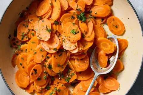 A close-up of a pot of glazed vichy carrots, glistening with butter and sprinkled with fresh parsley. A spoon rests in the pot, ready to serve.