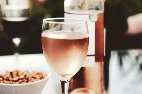 A glass of rosé wine, perhaps a Marsanne blend, sits on a table with a bowl of nuts and a bottle of wine.