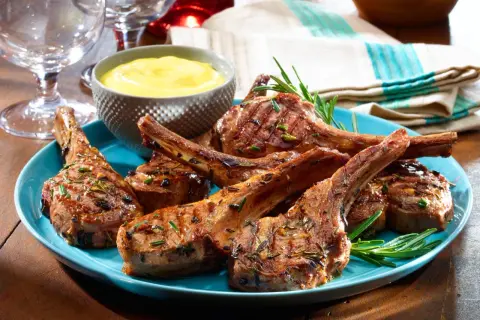 Grilled lamb chops, seasoned with rosemary, served on a blue plate with a side of yellow sauce. A taste of Spanish lamb meat.