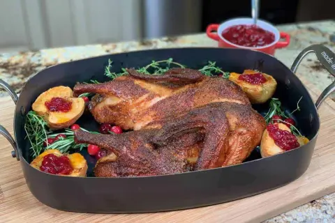 A beautifully roasted smoked duck rests in a dark roasting pan, garnished with fresh rosemary, cranberries, and baked apple halves topped with cranberry sauce.