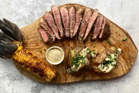 Juicy pork presa, grilled corn, and loaded baked potatoes served on a wooden board.