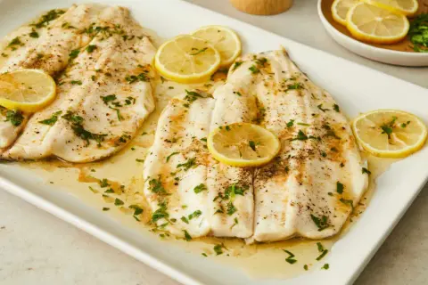Two beautifully cooked truchas fish fillets, glistening with butter and herbs, are garnished with fresh lemon slices on a white platter.