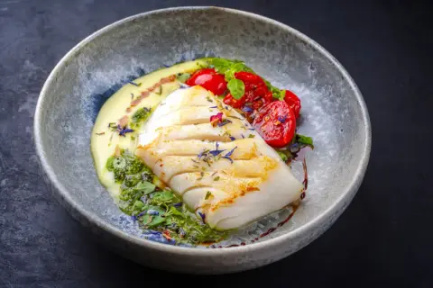 A beautifully plated poached fish recipe, featuring a white fish fillet with grilled marks, served with creamy sauce, pesto, and cherry tomatoes.