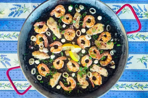 A delicious squid ink rice paella, brimming with plump shrimp, tender calamari rings, and flaky white fish, all garnished with fresh green onions and lemon wedges.