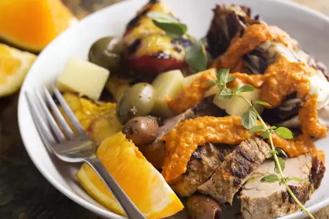 Slices of grilled spanish pork tenderloin with olives, orange wedges, and a vibrant romesco sauce.